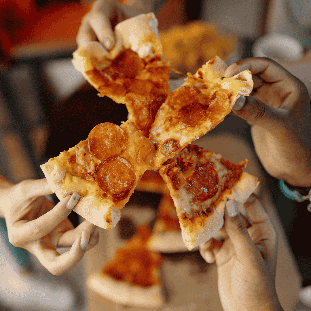 Four hands each holding a slice of pizza with pepperoni, with a blurred pizza box and pizza in the background.